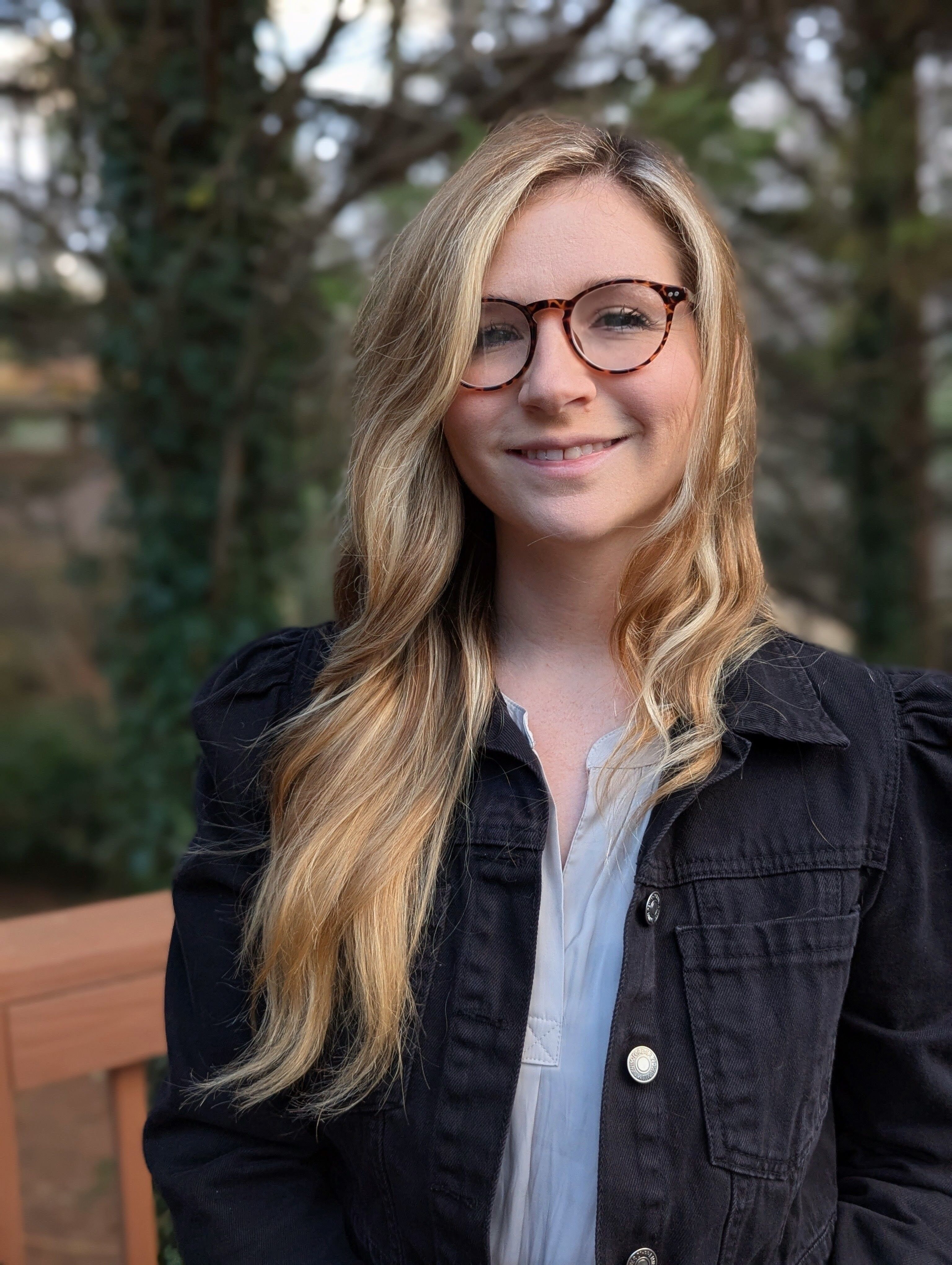 The image shows a young woman with blonde hair and glasses, smiling at the camera. She is wearing a denim jacket over a light-colored shirt. The background appears to be outdoors, with trees and foliage visible, suggesting a natural setting. The lighting is soft, highlighting her features.