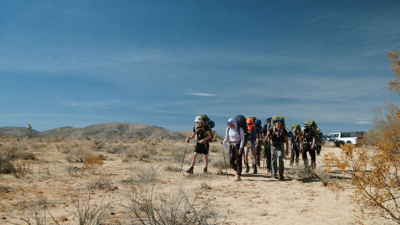 A group of people are hiking through a desert landscape. They are wearing backpacks and using trekking poles. The sky is blue and there are some mountains in the distance. A white pickup truck is parked on the right side of the image. The vegetation is sparse and dry.