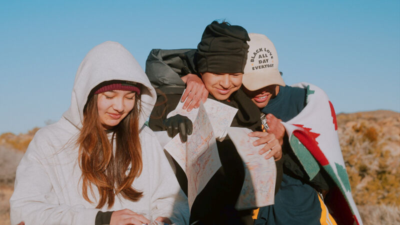 The image shows three young people outdoors, possibly hiking or exploring. One person, on the left, is wearing a white hooded jacket. The person in the middle is wearing a black beanie and dark clothing, holding a map. The third person, on the right, is wearing a baseball cap and is wrapped in a blanket with red and green stripes. They appear to be looking at the map together.
