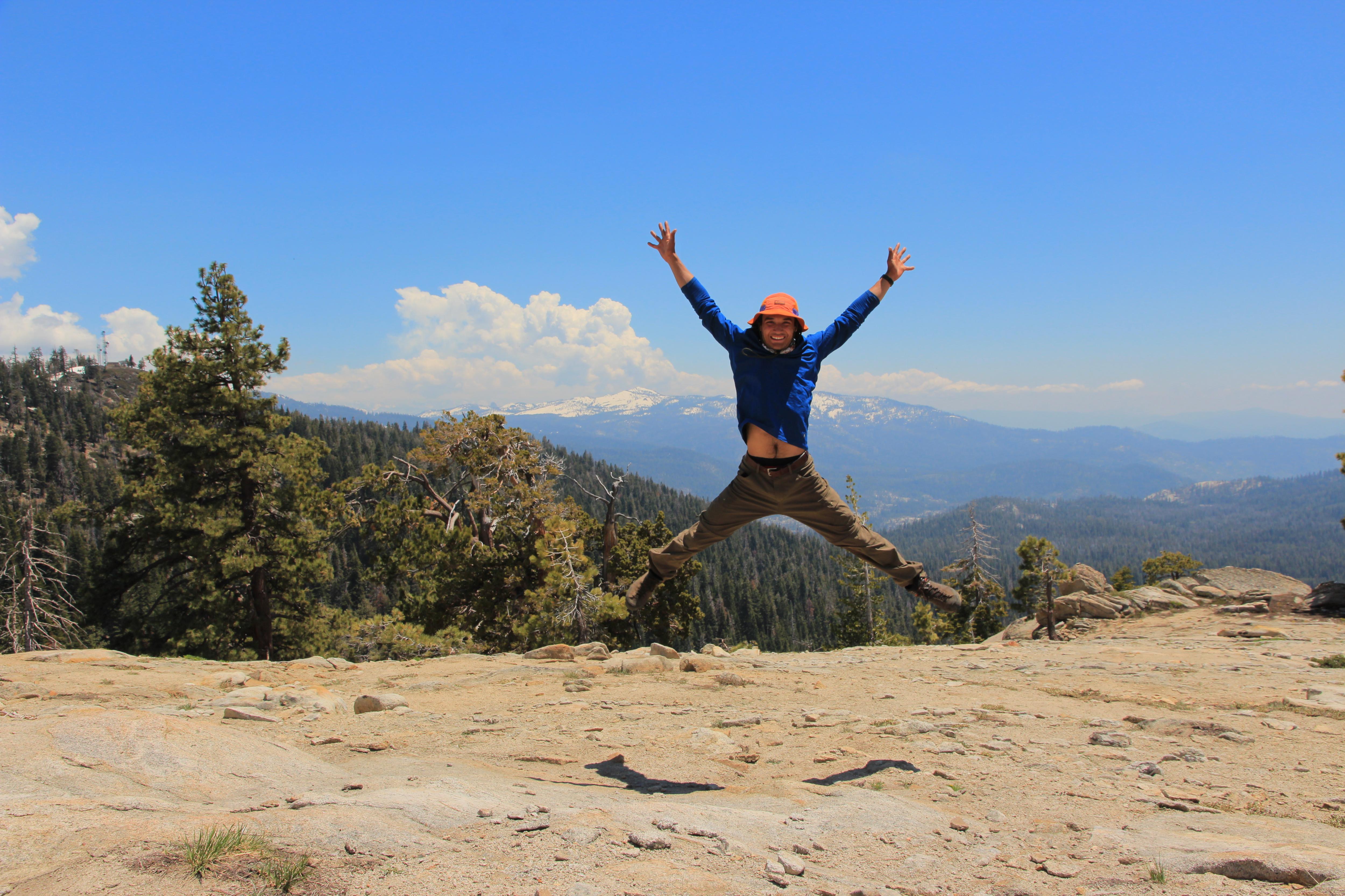 A person wearing a blue long-sleeved shirt, brown pants, and an orange hat is jumping in the air with their arms and legs spread out. They are on a rocky, sandy surface with trees and mountains in the background. The sky is blue with some clouds. The person appears to be enjoying themselves and celebrating.