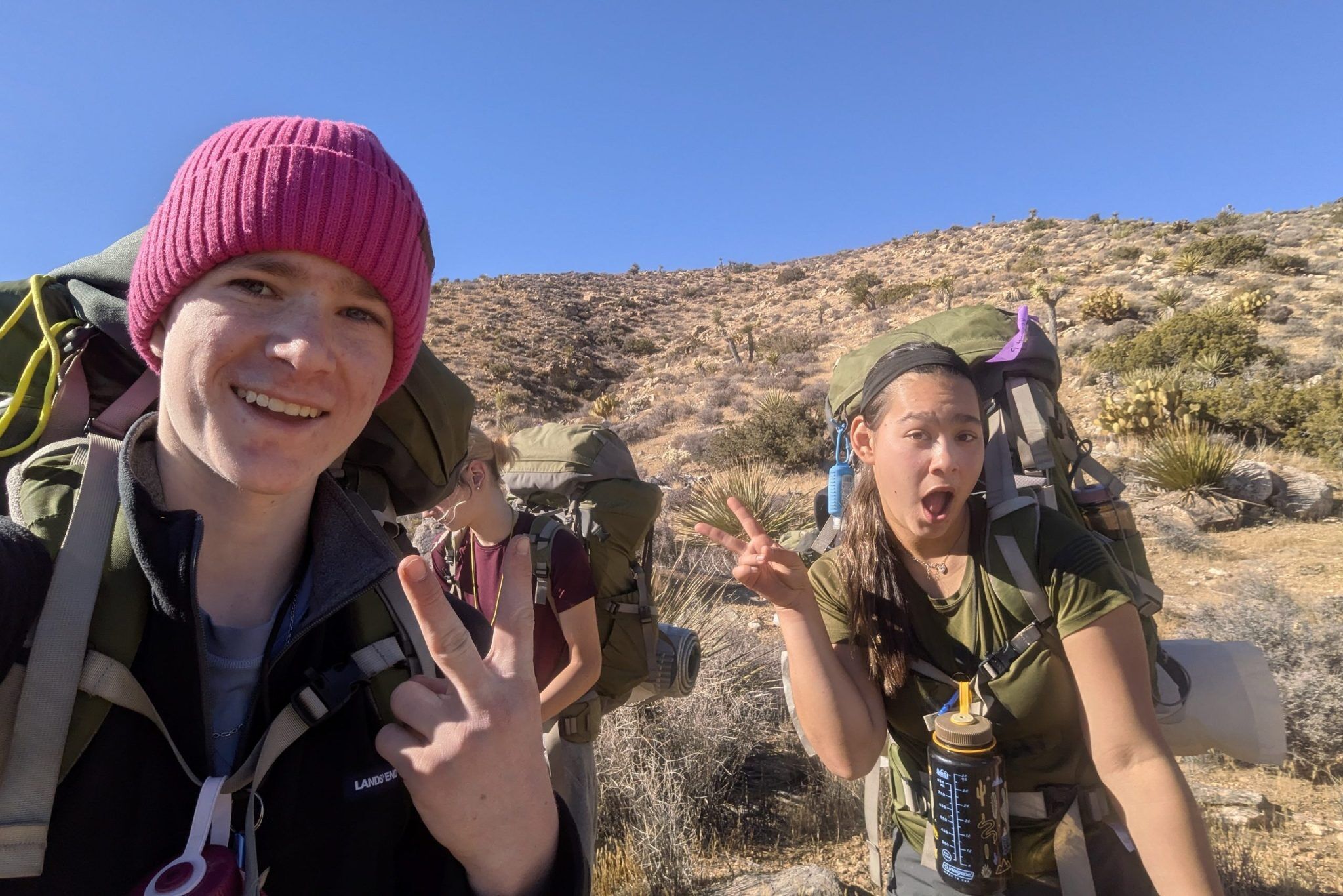 The image shows three young people hiking on a sunny day. They are wearing backpacks and appear to be on a trail in a desert-like environment. The person in the foreground on the left is smiling and holding up a peace sign. The person on the right is making a surprised face. The background shows a hill and a clear blue sky.
