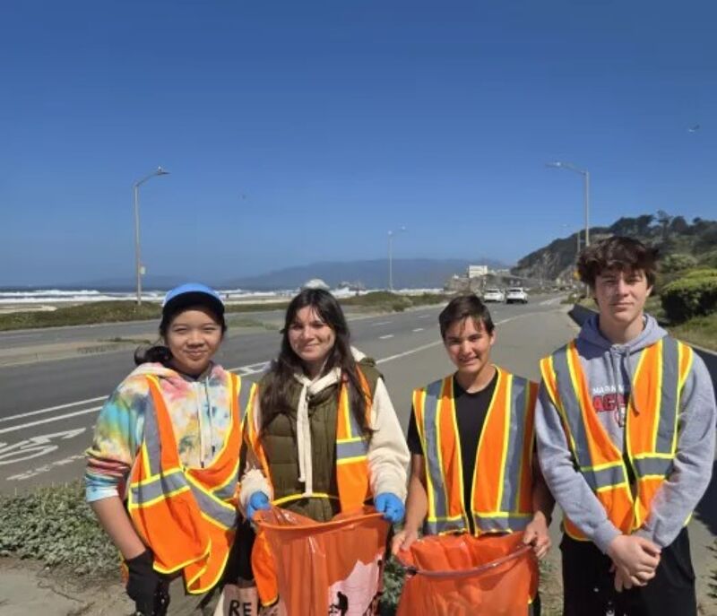 The image shows four young people wearing safety vests and holding orange trash bags. They appear to be participating in a cleanup activity, possibly along a road near the coast. The background includes a road, the ocean, and some buildings.