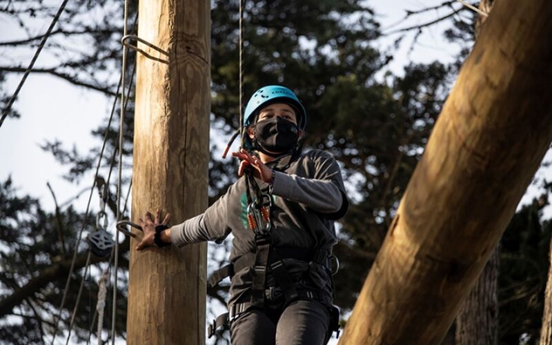 The image shows a person wearing a helmet and a face mask, navigating an obstacle course. They are attached to safety ropes and harnesses, indicating an adventure activity. The person is positioned between wooden poles, suggesting an outdoor setting, possibly a treetop adventure park.