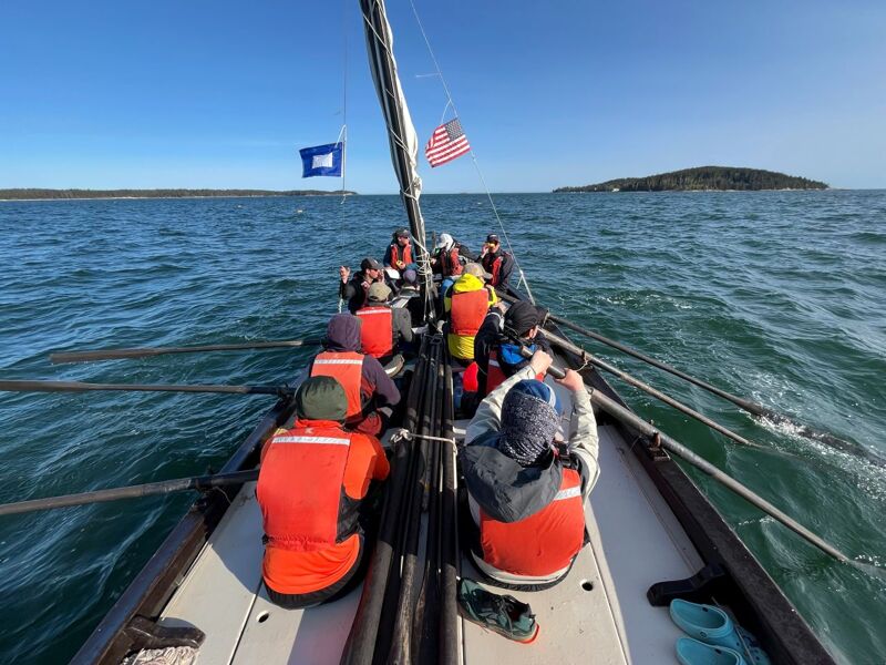 The image shows a group of people rowing a boat on a sunny day. They are wearing life jackets and appear to be working together. The boat is in the middle of the water, and there is land visible in the distance. The sky is blue and clear.