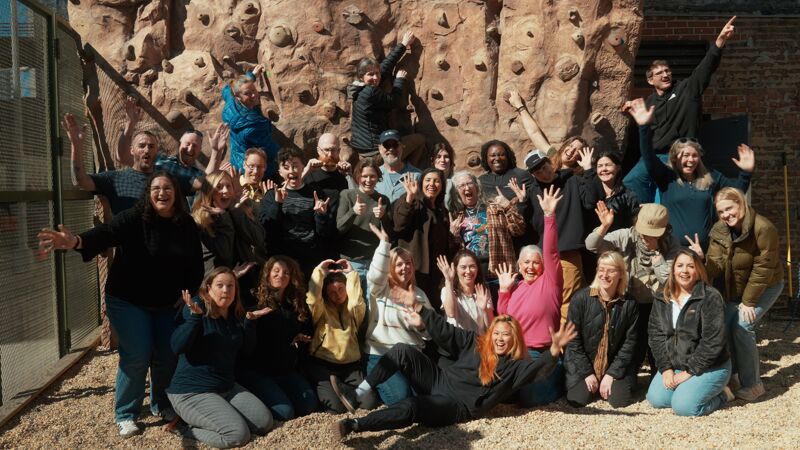 A large group of people are gathered outdoors, posing for a photo in front of a climbing wall. The group consists of men and women of varying ages, most of whom are smiling and looking at the camera. Some individuals are making hand gestures. The setting appears to be a recreational or team-building environment, with a brick building visible in the background.