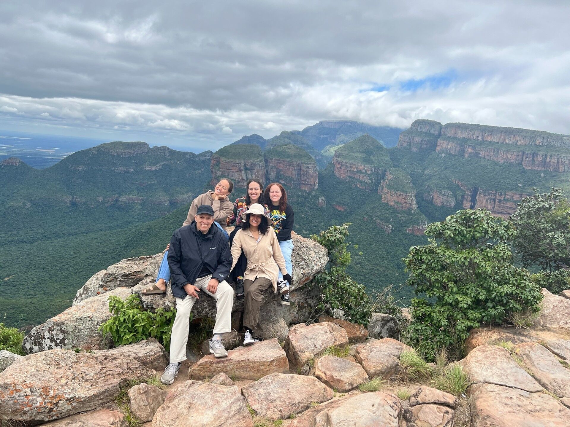 A group of five people is posing for a photo on a rocky outcrop, with a breathtaking view of lush green mountains and valleys under a cloudy sky. The people are dressed casually, and they appear to be enjoying a scenic hike or a visit to a viewpoint. The landscape suggests a natural park or a mountainous region, possibly in South Africa given the distinctive rock formations.