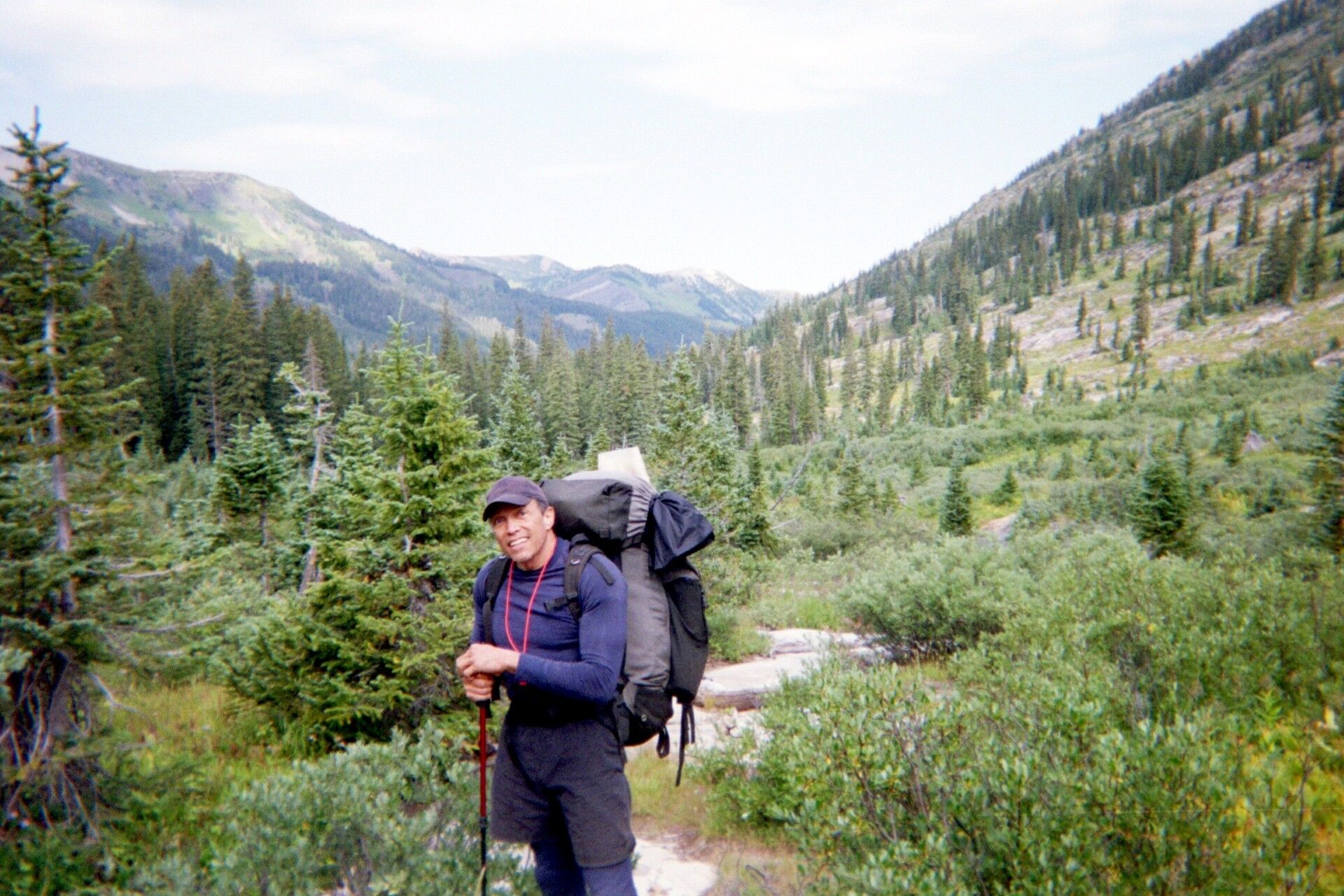 A man with a large backpack and a hiking pole is standing on a trail in a mountainous, forested area. He is wearing a blue long-sleeved shirt, dark shorts, and a baseball cap. The trail appears to be made of dirt and rocks, and it winds through dense green foliage and tall evergreen trees. The background shows rolling hills covered in trees, with some patches of lighter vegetation and a clear sky above. The overall scene depicts a scenic hiking or backpacking adventure in a natural, wilderness setting.