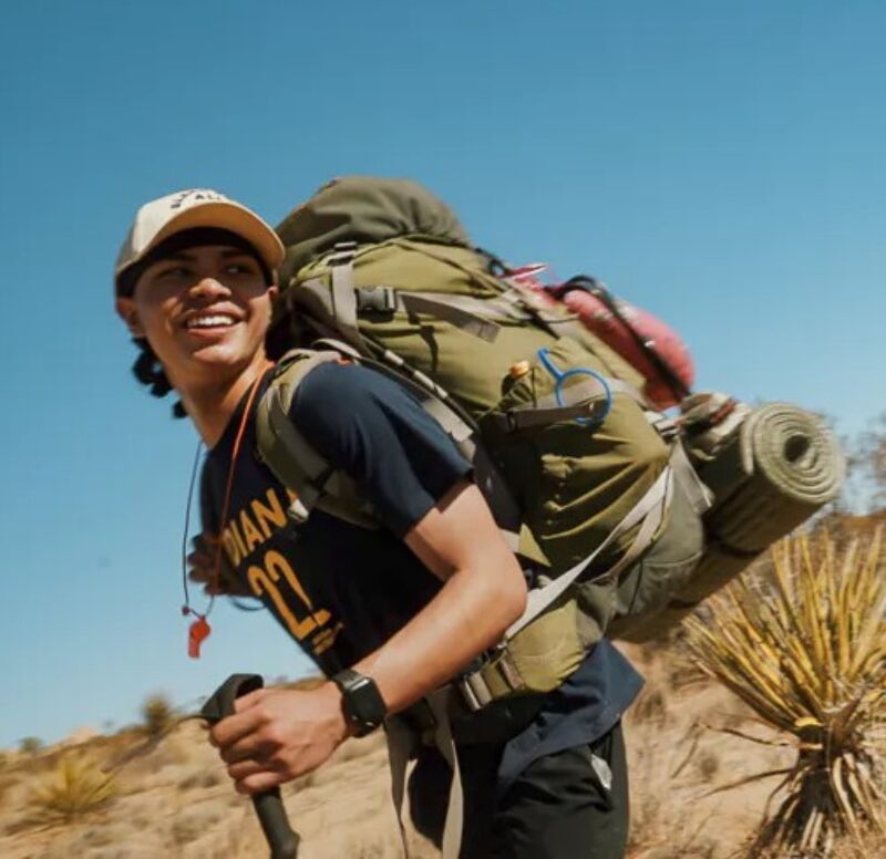 A young man is hiking in a desert landscape. He is wearing a baseball cap, a t-shirt, and a large backpack. He is holding a hiking pole in his right hand and smiling. The background consists of dry vegetation and a clear blue sky. The image suggests an outdoor adventure or backpacking trip.