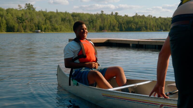 A young man wearing a white t-shirt and an orange life vest is sitting in a canoe on a lake. He is smiling and appears to be enjoying himself. The canoe is in the water near a wooden dock. In the background, there is a lush green forest. Another person is partially visible on the right side of the frame, near the canoe.