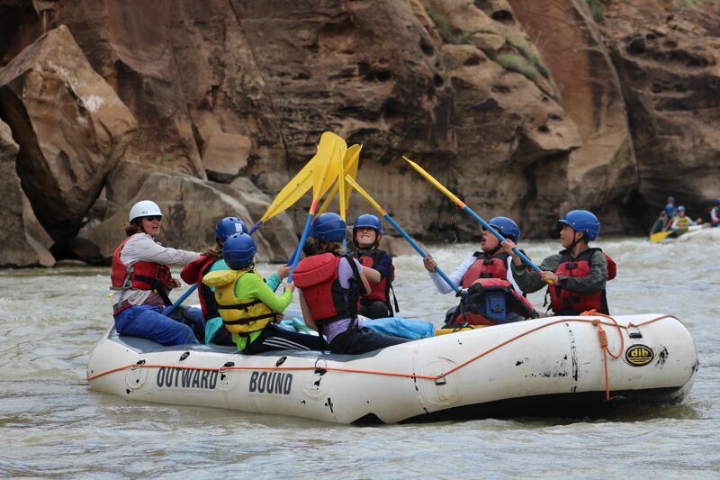 A group of people are enjoying a whitewater rafting trip. They are in an inflatable raft, paddling through a river with rocky cliffs on either side. Everyone is wearing helmets and life jackets for safety. The raft has the words "OUTWARD BOUND" printed on its side. The atmosphere appears adventurous and fun.