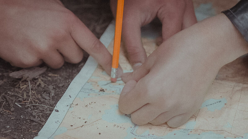 The image shows three hands pointing at a map with a pencil marking a specific location. The map is spread out on what appears to be the ground, possibly outdoors, given the presence of leaves and soil. The hands seem to be coordinating or planning a route, using the map as a guide. The focus is on the collaborative effort of navigation and decision-making.