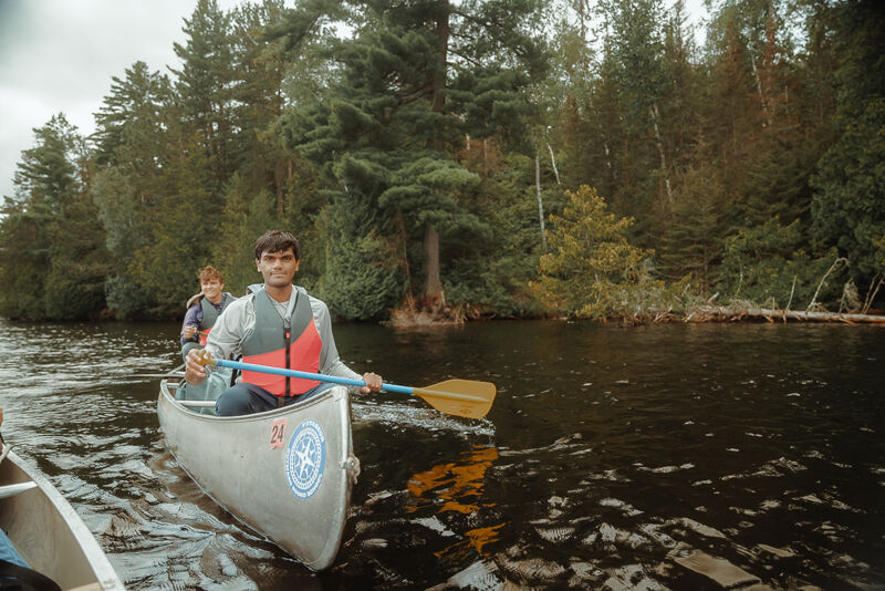 A canoe with two people is on a lake surrounded by trees. The person in the front is paddling and wearing a life vest. The water is dark and reflects the trees. The sky is overcast. The canoe has a number and a logo on its side.