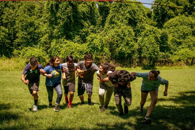 A group of people are running together in a grassy field, possibly participating in a team-building exercise or a game. They are holding onto each other, suggesting they are working together towards a common goal. The background features lush green trees, indicating the event is taking place outdoors on a sunny day.