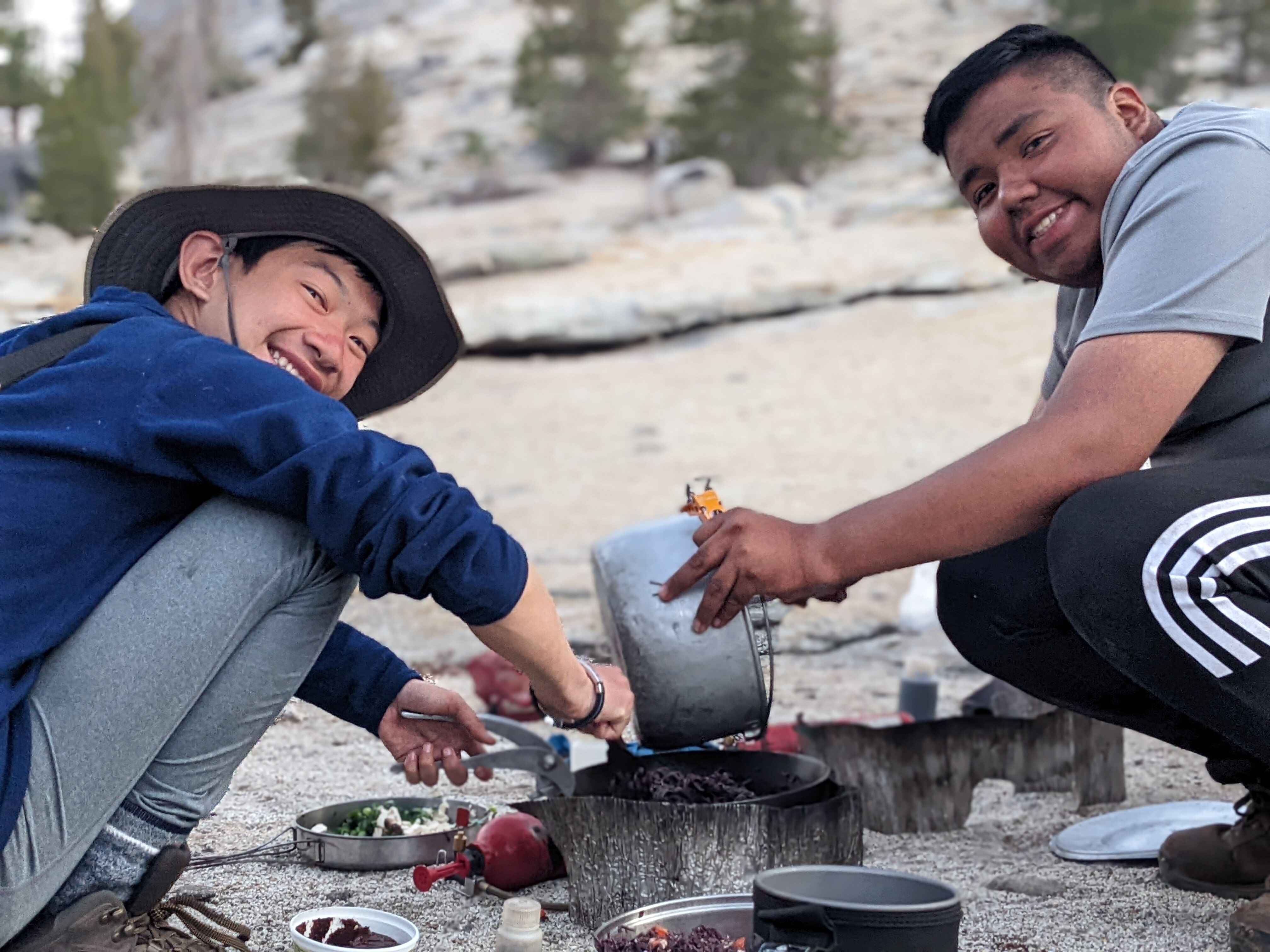 Two young men are outdoors, possibly camping. One on the left wears a hat and blue jacket, holding a container over a cooking pot. The other, on the right, wears a gray shirt and black pants with a white stripe. They appear to be preparing food, with cooking equipment and ingredients visible around them. The background shows a natural setting with trees and rocks.
