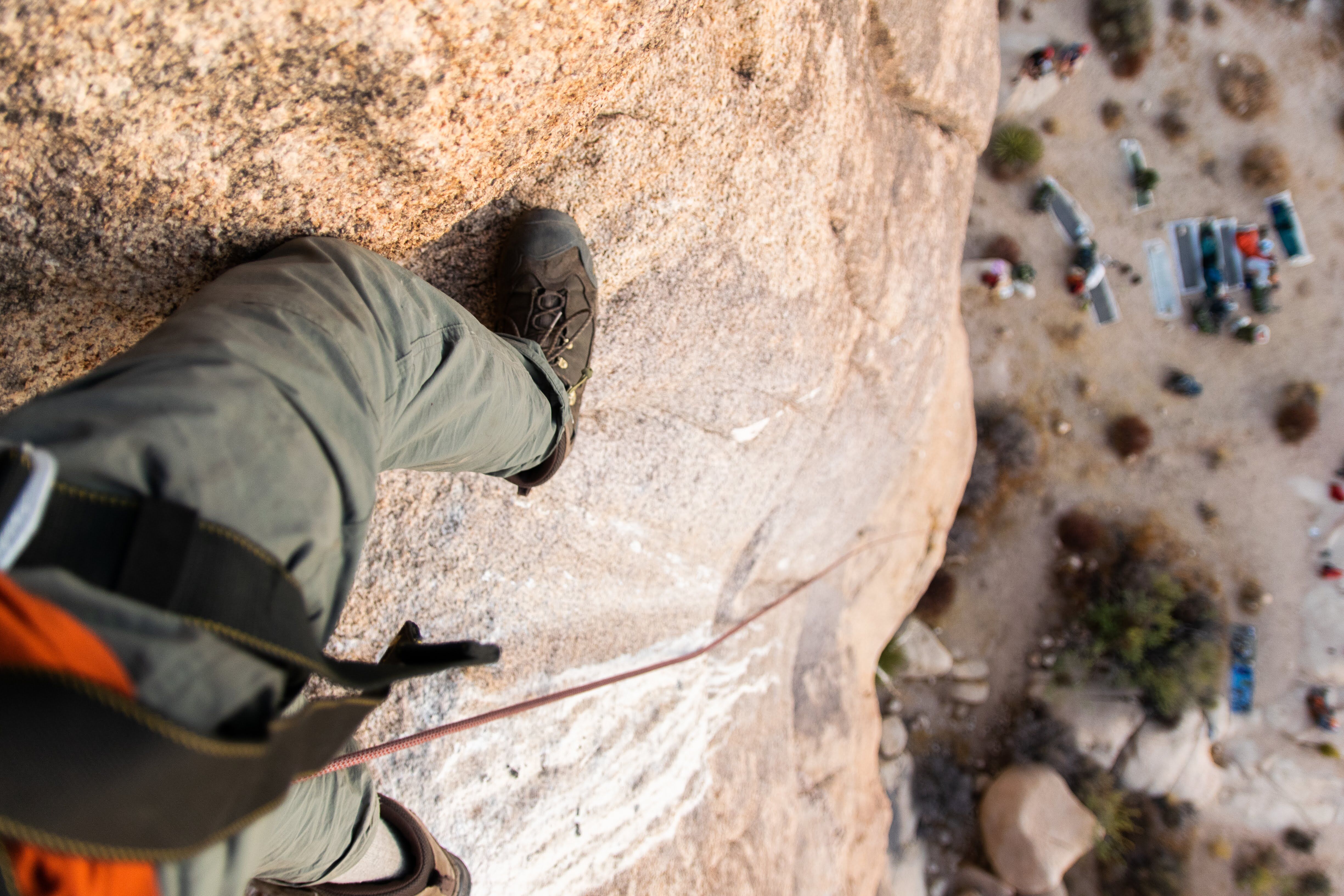 The image shows a first-person perspective of a rock climber scaling a cliff face. The climber is wearing climbing shoes, pants, and a harness. A rope is visible, suggesting they are using safety equipment. In the background, there's a view of the ground below, possibly a campsite or a natural landscape with sparse vegetation. The angle emphasizes the height and the challenge of the climb.