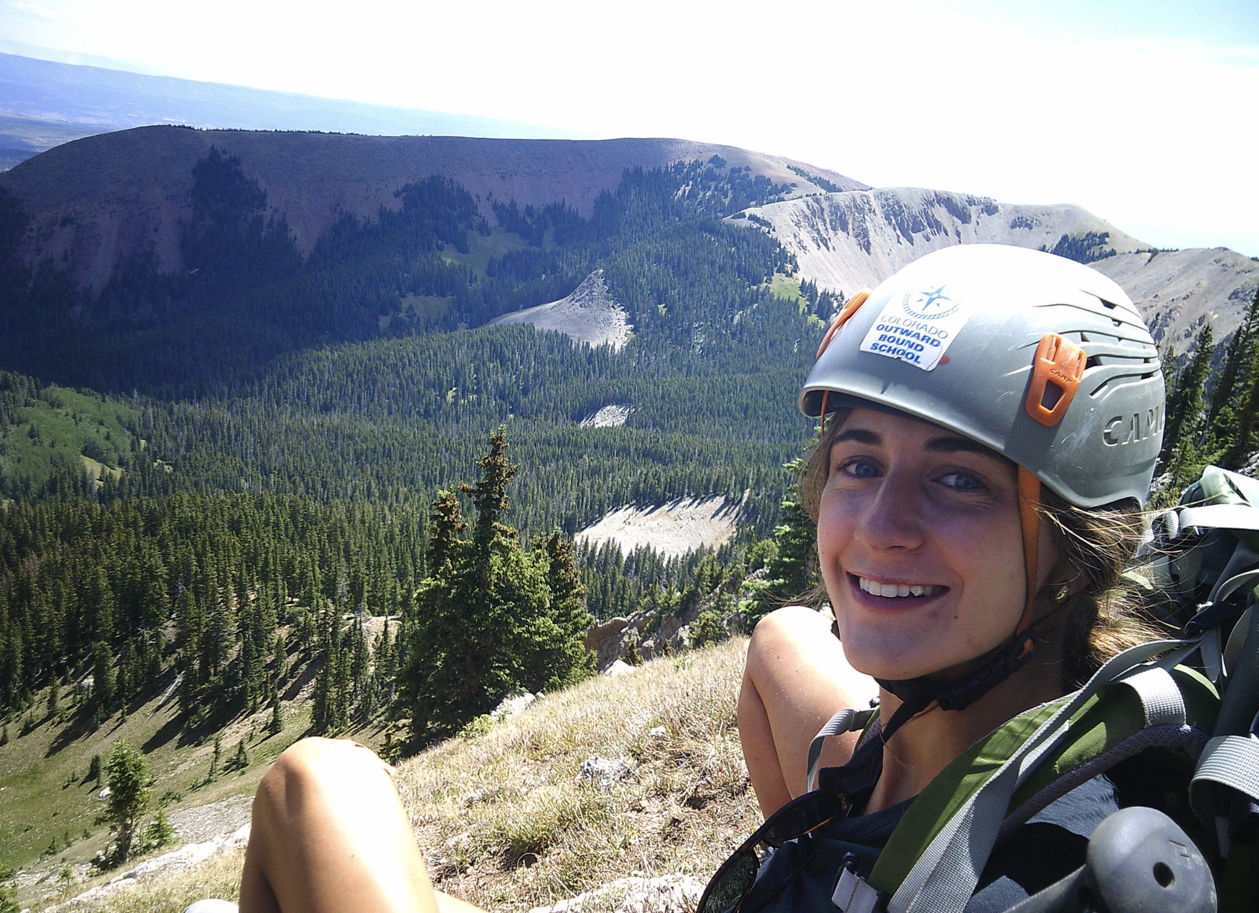 A young woman with a helmet and backpack smiles at the camera, sitting on a grassy slope with a vast, tree-covered mountain range and a clear blue sky in the background. She appears to be on a hiking or climbing expedition, enjoying the scenic vista.