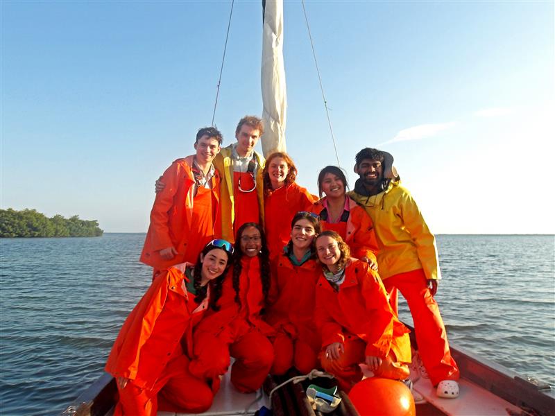 A group of nine young adults, dressed in bright orange raincoats and some in yellow, are posing for a photo on a sailboat. They are on the water, with a clear blue sky above and some greenery visible in the distance. The mood appears cheerful and adventurous as they enjoy a day out on the sea.