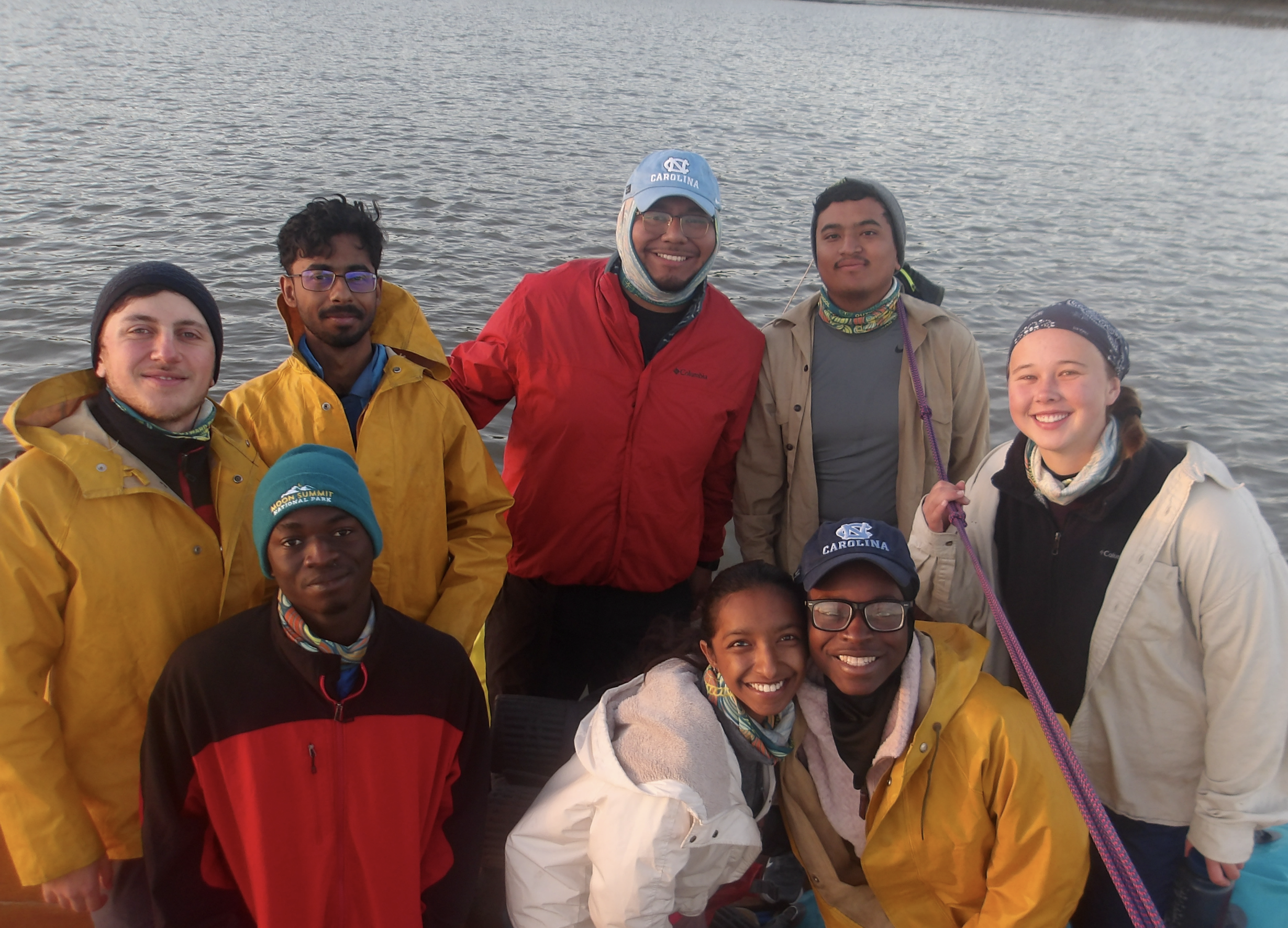 A group of nine young adults, appearing to be students, are posing for a photo on a body of water. Most are wearing yellow or tan rain jackets, and some have blue UNC logos on their hats or jackets. The group includes a diverse mix of ethnicities and genders, and they all seem to be in good spirits. The water behind them is calm, suggesting a peaceful setting.