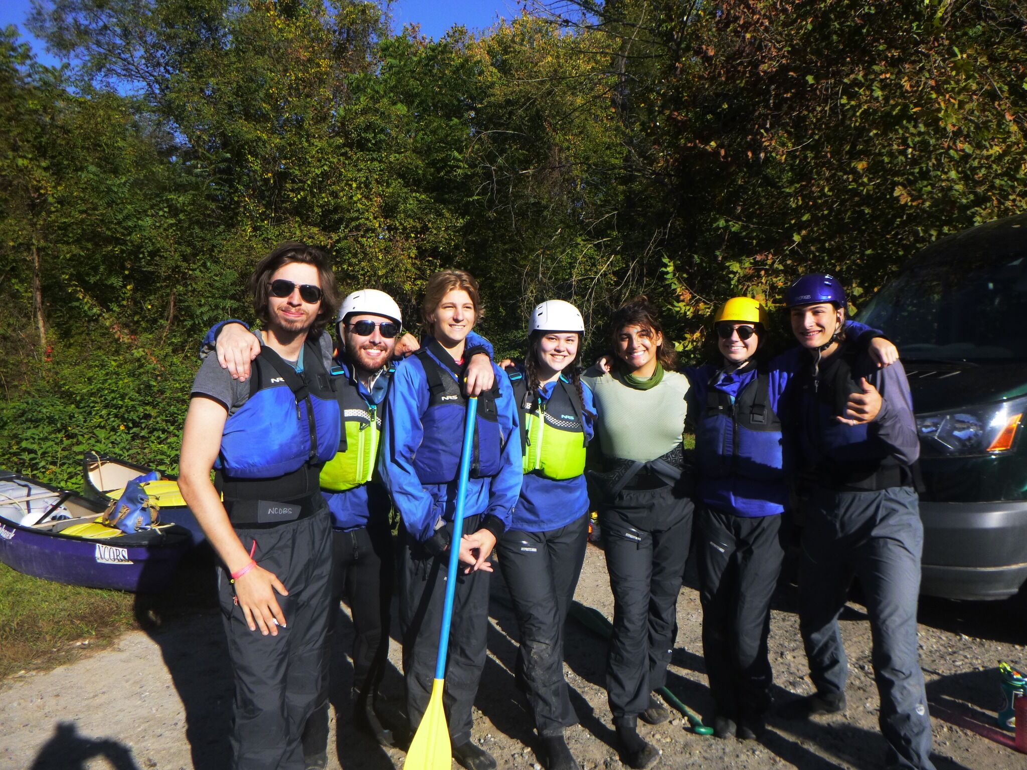 A group of seven people, dressed in life vests and helmets, are posing for a photo outdoors. They appear to be ready for a water activity like rafting or kayaking, with a raft and paddles visible. The weather is sunny, with trees in the background.