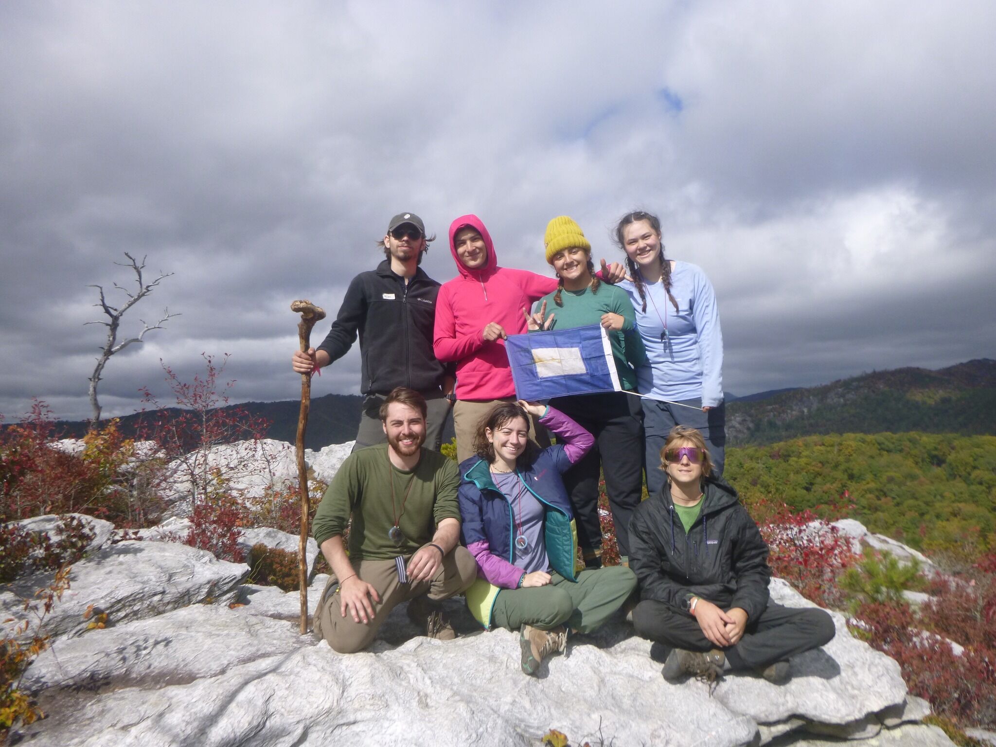 A group of seven people are posing for a photo on a rocky outcrop, with a scenic view of autumn foliage and mountains in the background. They are dressed in outdoor clothing, suggesting they are hikers or nature enthusiasts. One person holds a blue and white flag. The sky is partly cloudy, adding a dramatic effect to the landscape.