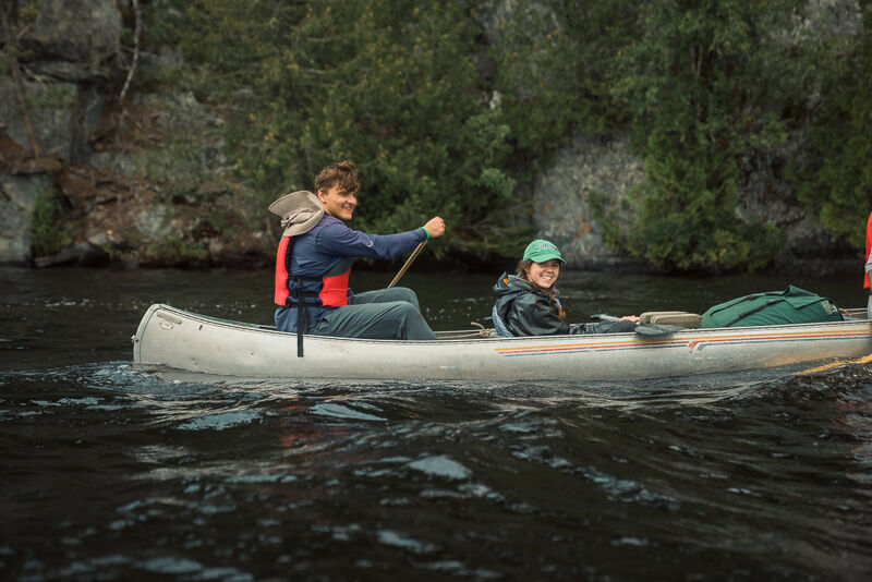 The image shows two people paddling a canoe on a body of water. The person in the front is wearing a blue shirt and an orange life vest and is actively paddling. The person in the back is wearing a green hat and a dark jacket. The canoe is moving through the water, creating ripples. The background features lush green trees and rocky cliffs. The overall scene suggests a peaceful outdoor activity.