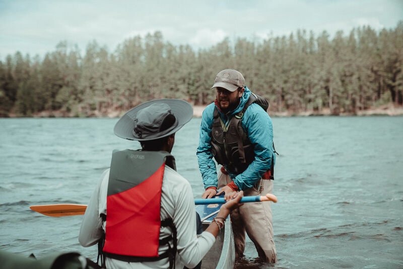 Two people are standing in the water near a forest. One person is wearing a wide-brimmed hat and a red life vest, while the other is wearing a baseball cap and a blue life vest. They appear to be holding onto a kayak or canoe. The water is calm, and the sky is overcast.
