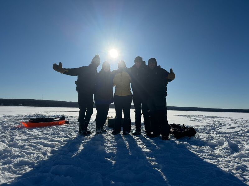 The image shows a group of six people standing on a snow-covered field, likely a frozen lake, with the sun shining brightly behind them. They are silhouetted against the bright light, and their poses suggest they are enjoying the sunny day. There's a sled on the left side of the image. The background features a distant treeline, indicating the edge of the snowy expanse.