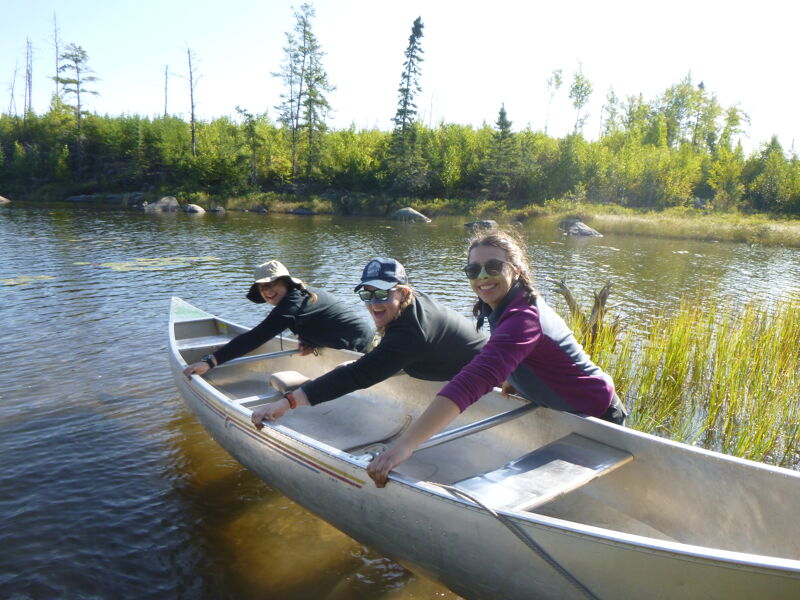 The image shows three people in a canoe on a lake. They are all smiling and appear to be enjoying themselves. The weather looks sunny and the scenery is beautiful. There are trees and grass in the background.