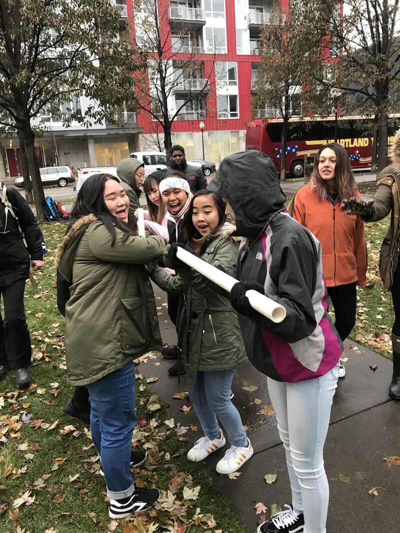The image shows a group of young people outdoors, seemingly engaged in a playful activity. They are gathered closely together, with some holding a white tube. The expressions on their faces suggest excitement and enjoyment. The background includes a red building, trees, and a bus, indicating an urban or suburban setting.