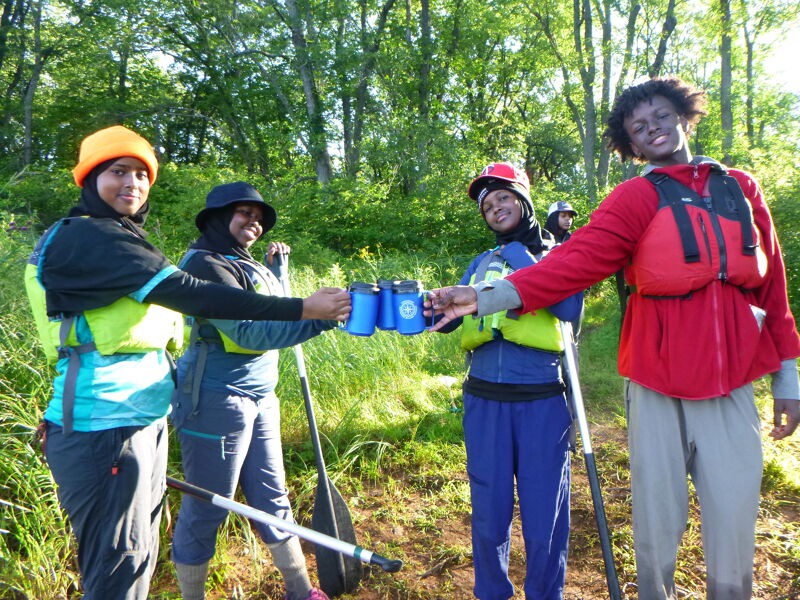 The image shows a group of people in outdoor gear, possibly near a body of water. They are holding up cans and appear to be celebrating or toasting. The people are wearing life vests and hats, suggesting they might be involved in water activities like kayaking or rafting. The background includes trees and greenery.
