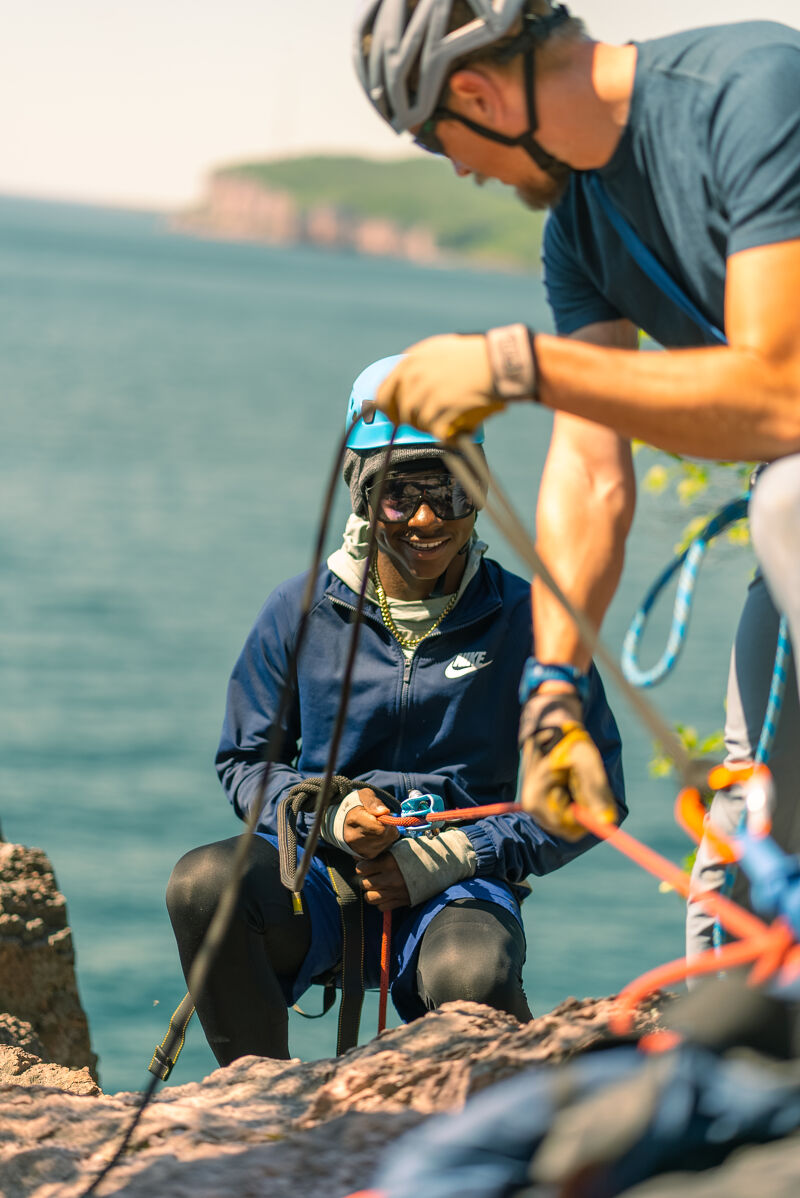A person wearing a blue jacket and helmet is sitting on a rocky cliff, seemingly preparing for or engaged in rock climbing or rappelling. Another person, also wearing a helmet, is standing nearby, possibly assisting or instructing. Ropes and climbing gear are visible, suggesting an outdoor adventure activity. The background features a body of water and a distant shoreline, indicating a scenic location for this activity.