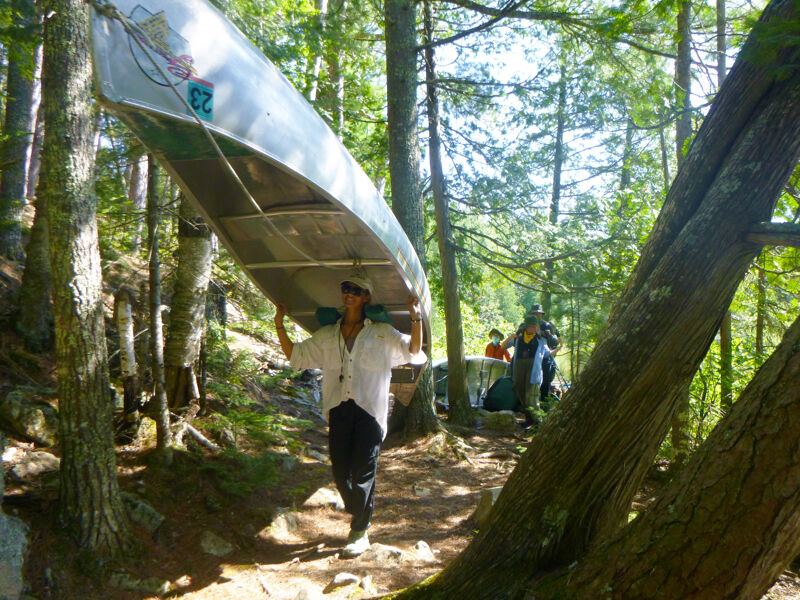 A person is carrying a canoe over their head while walking on a dirt path in a forest. The canoe is silver and appears to be lightweight. The person is wearing a light-colored shirt and dark pants. The path is surrounded by trees and foliage, and there are other people in the background. The scene suggests a portage during a canoeing or kayaking trip.