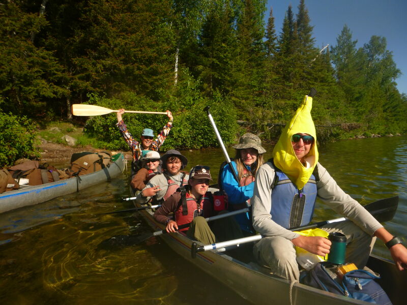 A group of people are enjoying a canoe trip on a lake surrounded by lush greenery. They are in two canoes, with one person in the front canoe wearing a banana hat and sunglasses. The others are wearing hats and life jackets. They are paddling and smiling, enjoying the scenery and the company. The sun is shining, and the water is calm.