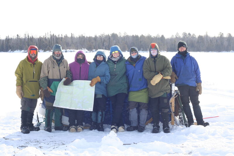 A group of eight people stands in the snow, dressed in winter clothing. They are holding a map and appear to be on an outdoor adventure. The background shows a snowy landscape with trees in the distance. The lighting suggests it is a bright, sunny day.