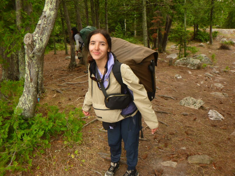A young woman is hiking in a forest, carrying a large backpack. She is wearing a light-colored jacket, blue pants, and hiking boots. The forest is dense with trees and foliage, and the ground is covered with leaves and dirt. The woman is smiling and looking at the camera. There is another person with a backpack in the background.