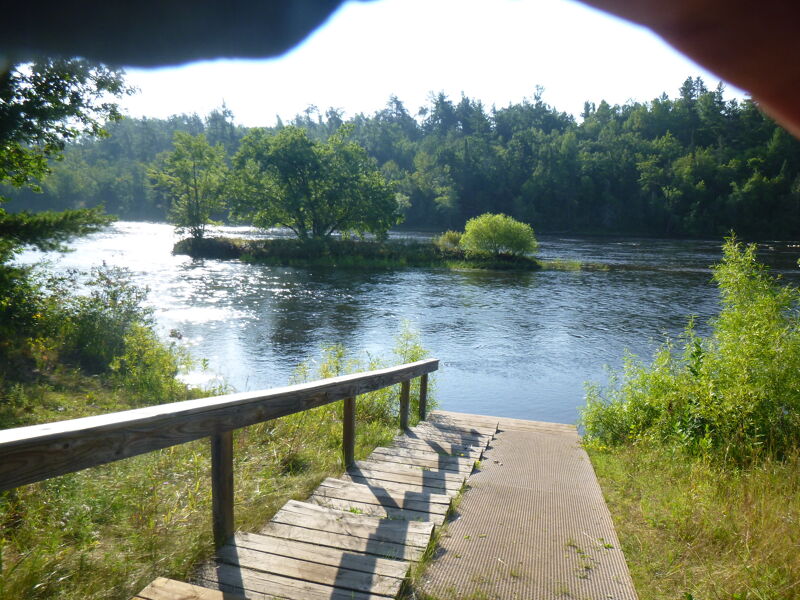 The image shows a tranquil river scene with a wooden staircase leading down to the water. Lush greenery lines the banks, and a small island sits in the middle of the river. The water reflects the sunlight, creating a shimmering effect. The scene evokes a sense of peace and natural beauty.