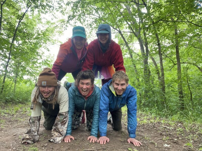 The image shows five people posing in a forest. Four people are on their hands and knees, while the fifth person is standing on top of them. They are all smiling and appear to be having fun. The background consists of trees and foliage.