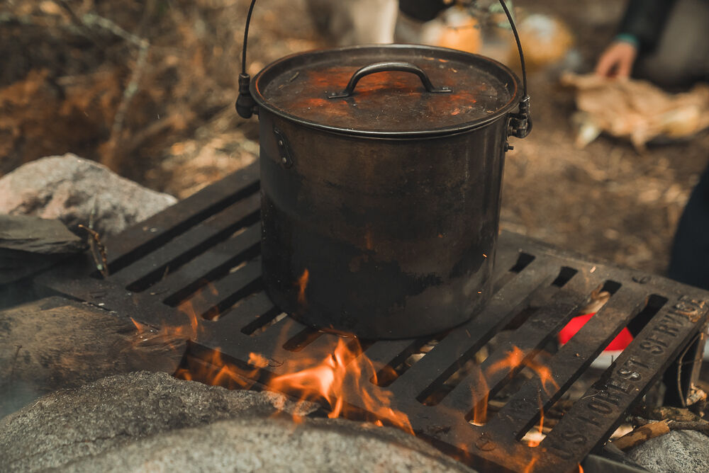 The image shows a metal pot with a lid sitting on a grate over a campfire. The pot appears to be used for cooking, and flames are visible beneath it, suggesting that something is being heated or cooked inside. The surrounding environment seems to be outdoors, possibly a campsite, with rocks and other natural elements visible.