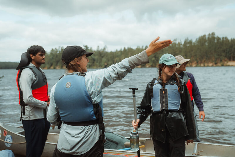 A group of four people are standing near a canoe on a cloudy day, seemingly preparing for a water activity. They are all wearing life jackets, and one person is gesturing towards the water, possibly giving instructions or pointing out a direction. The background features a lake and a treeline, suggesting they are in a natural, outdoor setting.