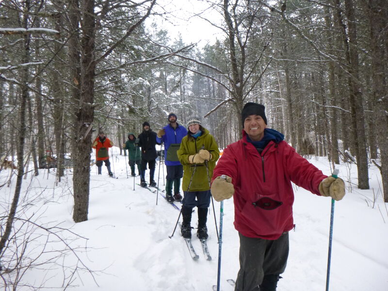 The image shows a group of people cross-country skiing through a snowy forest. They are all wearing winter clothing, including jackets, hats, and gloves. The person in the foreground is smiling at the camera. The trees are bare, and the sky is overcast.