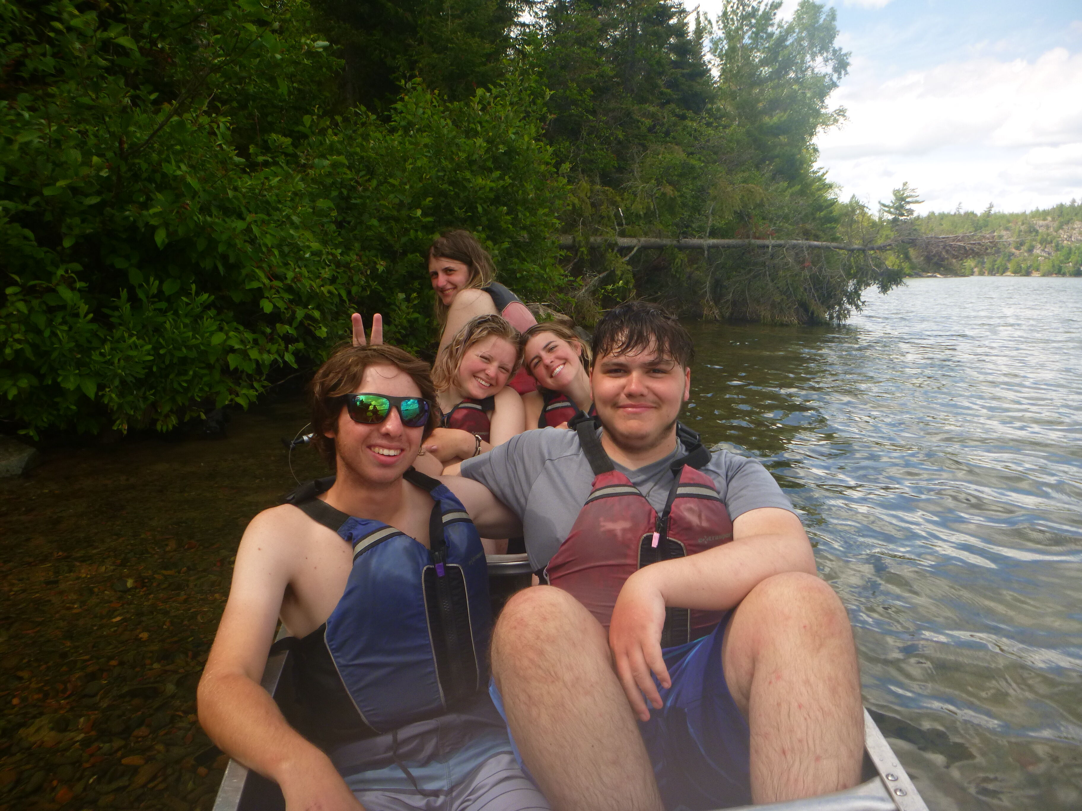 A group of five people are in a canoe on a lake. The person in the front is wearing sunglasses and making a peace sign with their fingers. Everyone in the canoe is wearing a life jacket. The background shows trees and the lake extending into the distance under a partly cloudy sky.
