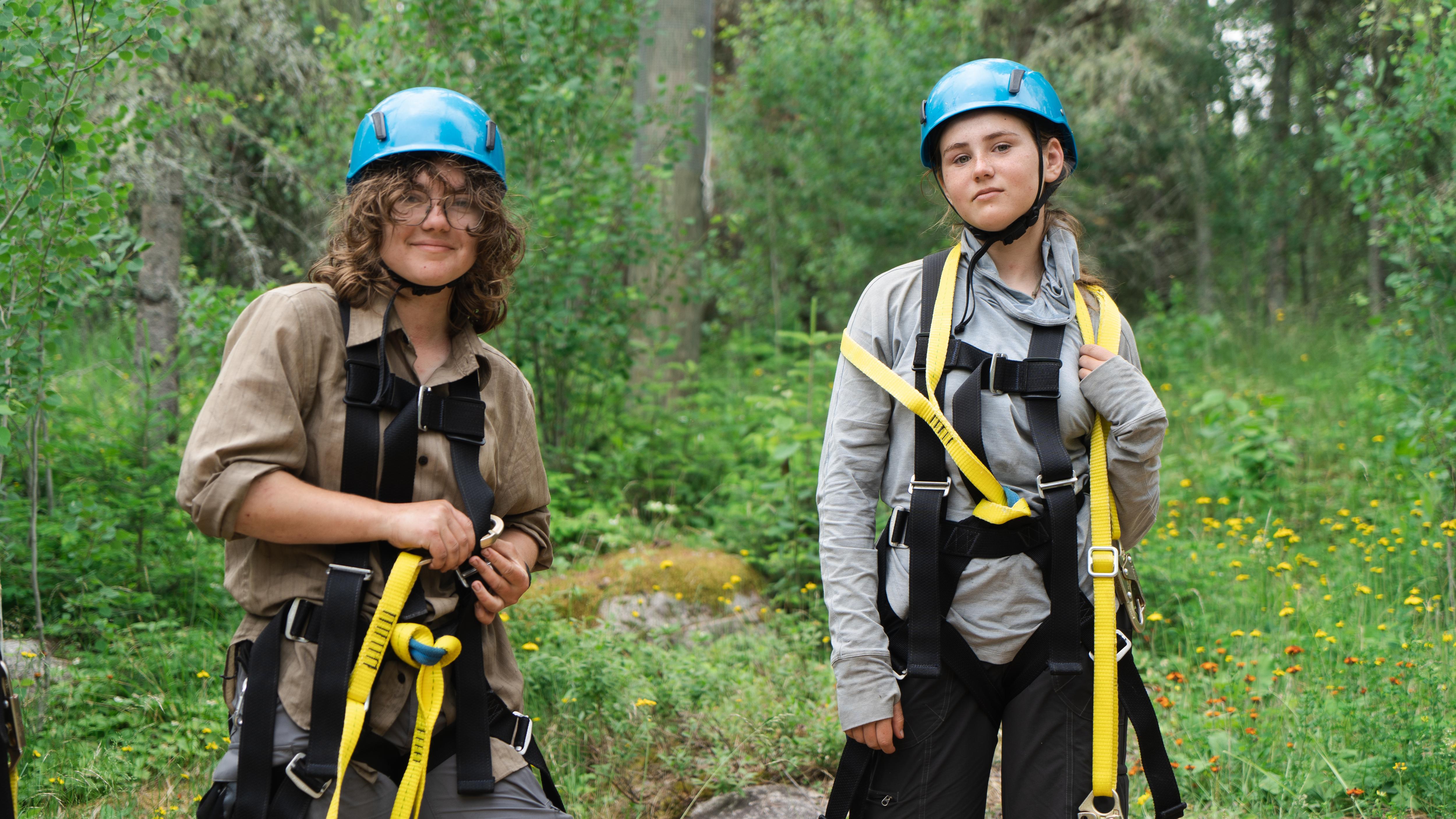 Two young women stand in a forest, geared up for a zip-lining adventure. They both wear helmets and harnesses, ready to take on the trees. The woman on the left has curly hair and a khaki shirt, while the woman on the right sports a gray top. Sunlight filters through the trees, creating a dappled effect on the forest floor.