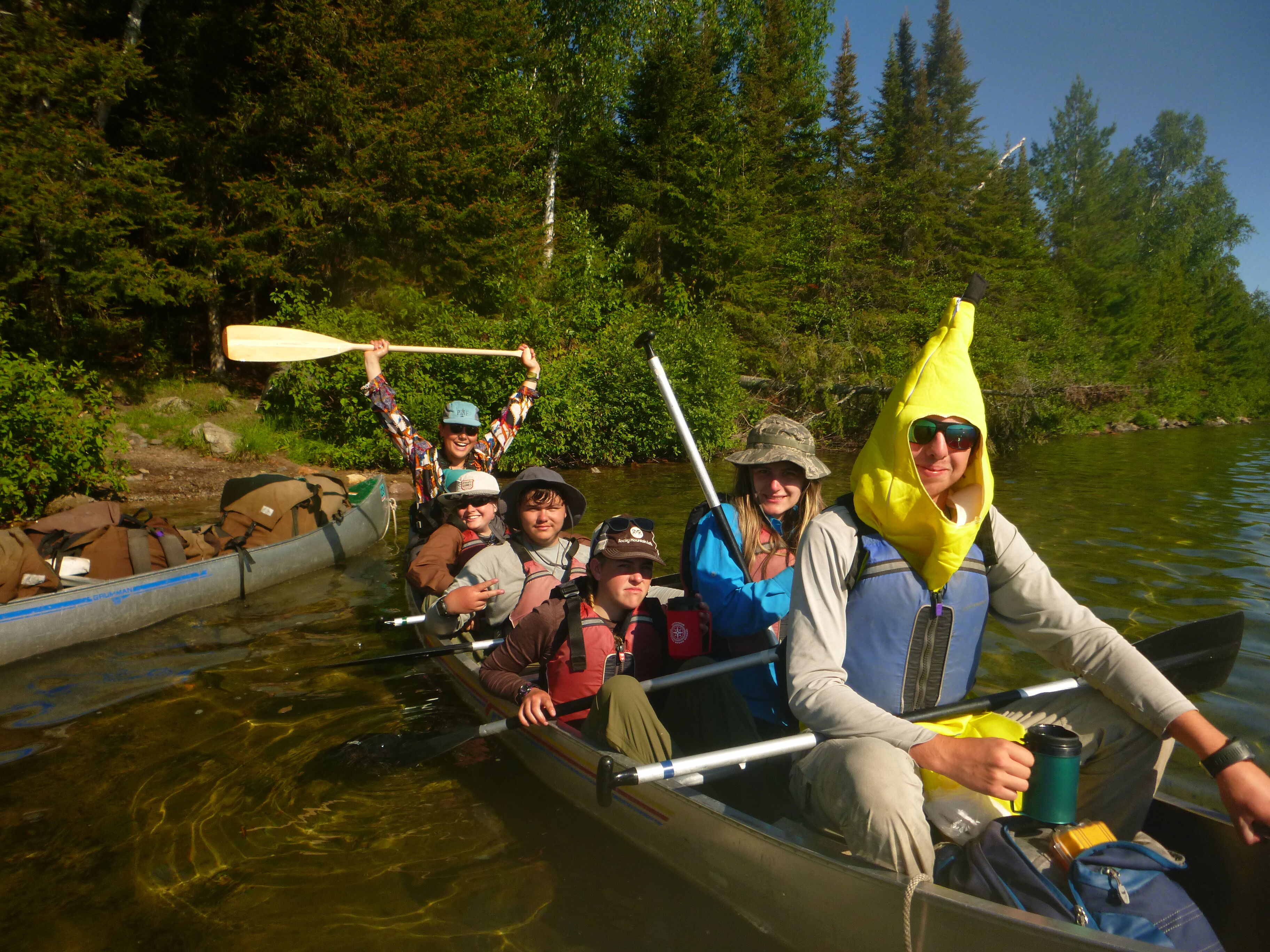 A group of people are enjoying a canoe trip on a sunny day. They are paddling through a calm lake surrounded by lush green trees. One person is wearing a banana suit. The canoes are loaded with gear, suggesting a multi-day adventure. Everyone seems to be in good spirits, enjoying the beautiful scenery and the company of friends.
