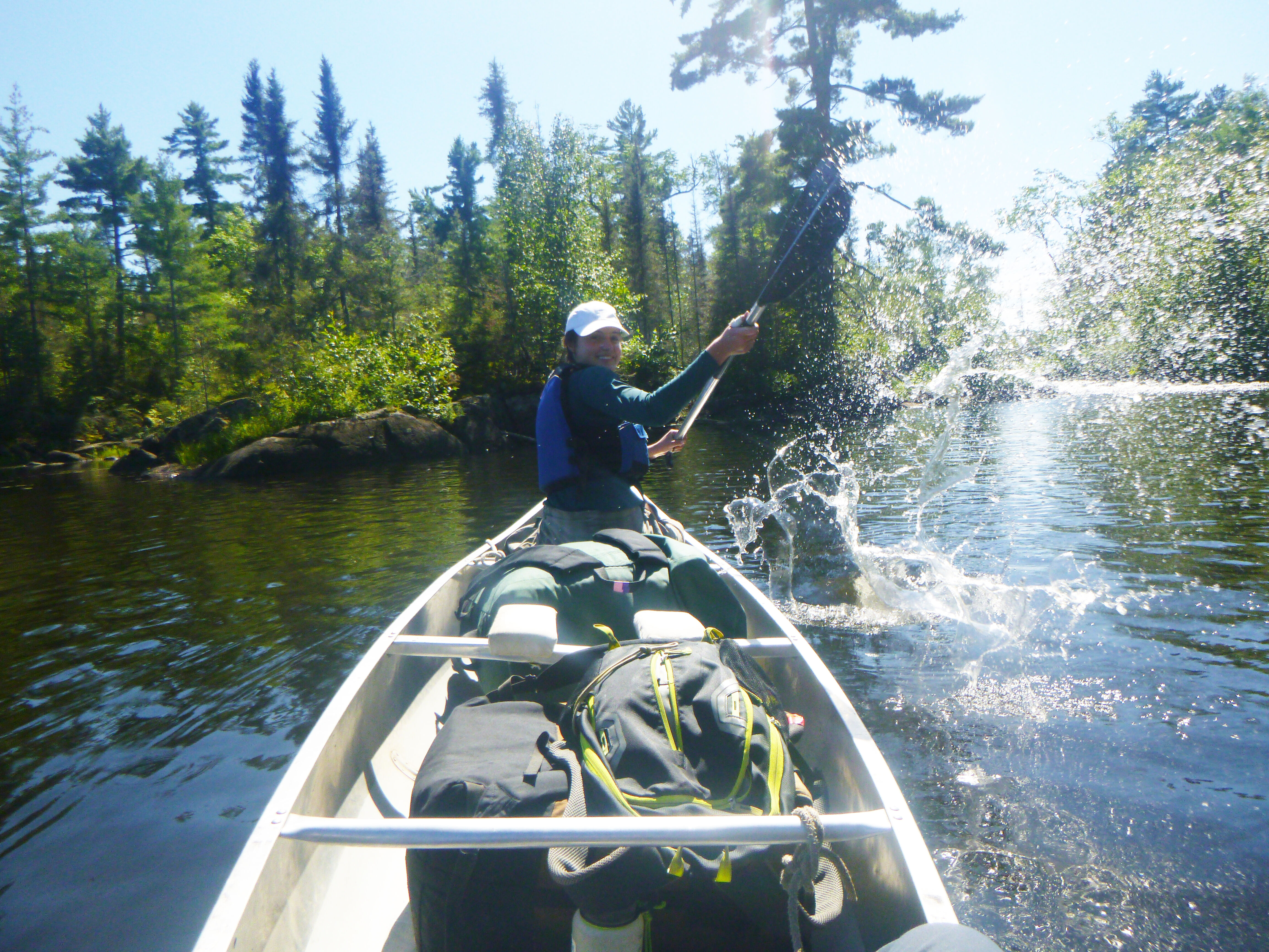 A person in a canoe is fishing. The person is wearing a hat and a life jacket. The canoe is in a lake or river, and there are trees in the background. The person has caught a fish, and water is splashing around the canoe.