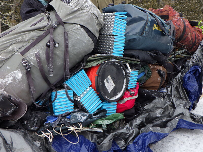 A pile of camping gear sits on the ground, possibly covered in light snow. Backpacks in shades of gray and blue are prominent, along with rolled-up blue sleeping pads. There are also various other items like buckets, ropes, and colorful bags mixed in, creating a cluttered scene of outdoor equipment ready for an adventure.
